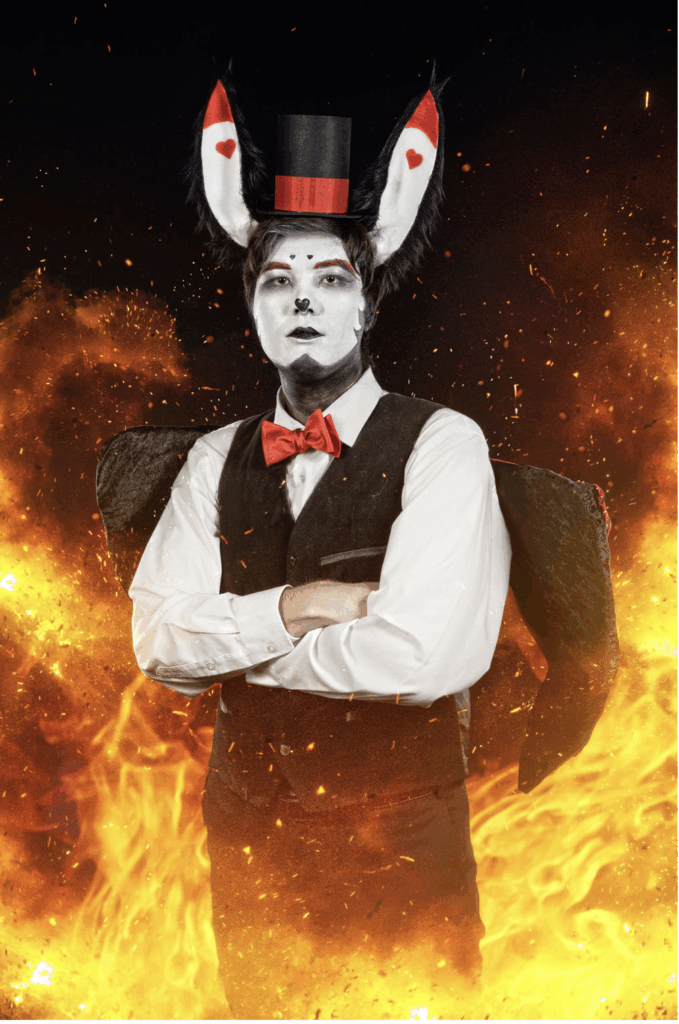 Caleb stands in cosplay confidently with arms crossed, dressed in an elaborate costume featuring black rabbit ears with red hearts, a small top hat with red trim, a red bowtie, and a black vest over a white shirt. Their face is painted silver with black and red makeup resembling a playing card theme. They are surrounded by dramatic flames and glowing embers, creating a powerful, infernal atmosphere.