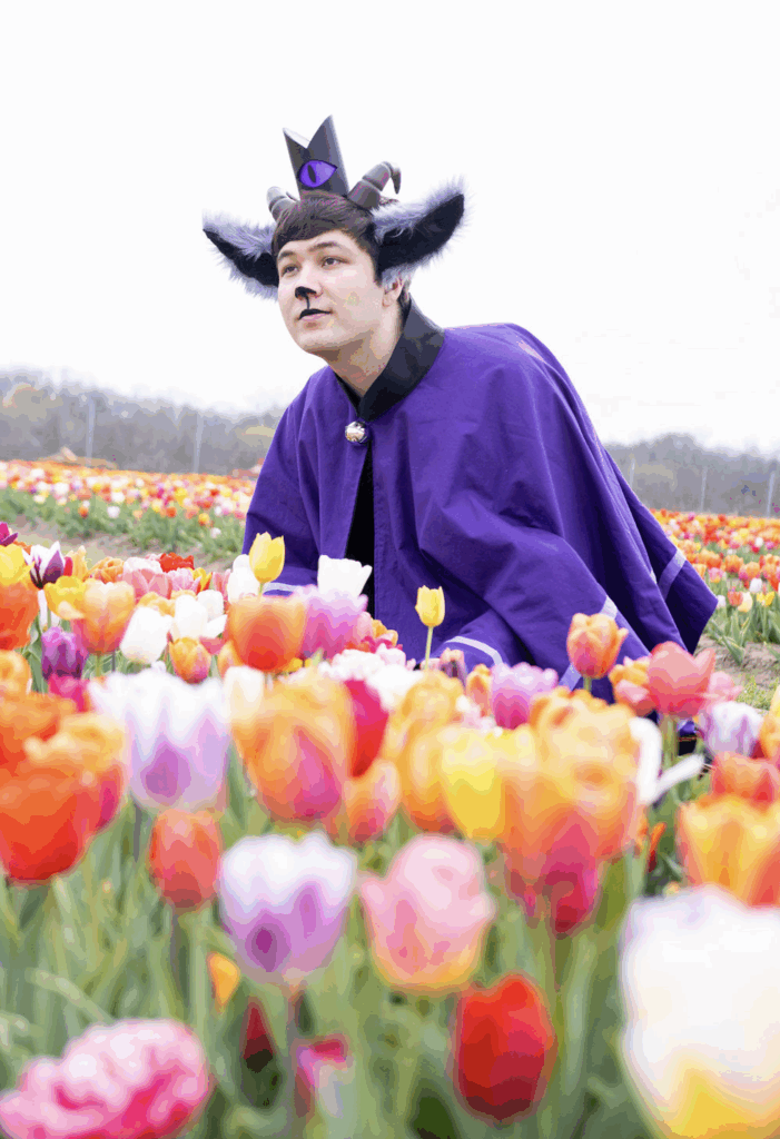 Caleb in a whimsical costume crouches among a vibrant field of multicolored tulips. They wear a purple cloak with a gemstone clasp, a black and purple crown with a single large eye motif, and large black-and-white animal ears accented by horns. Their makeup and expression evoke a playful or curious demeanor, contrasting with the serene natural background.