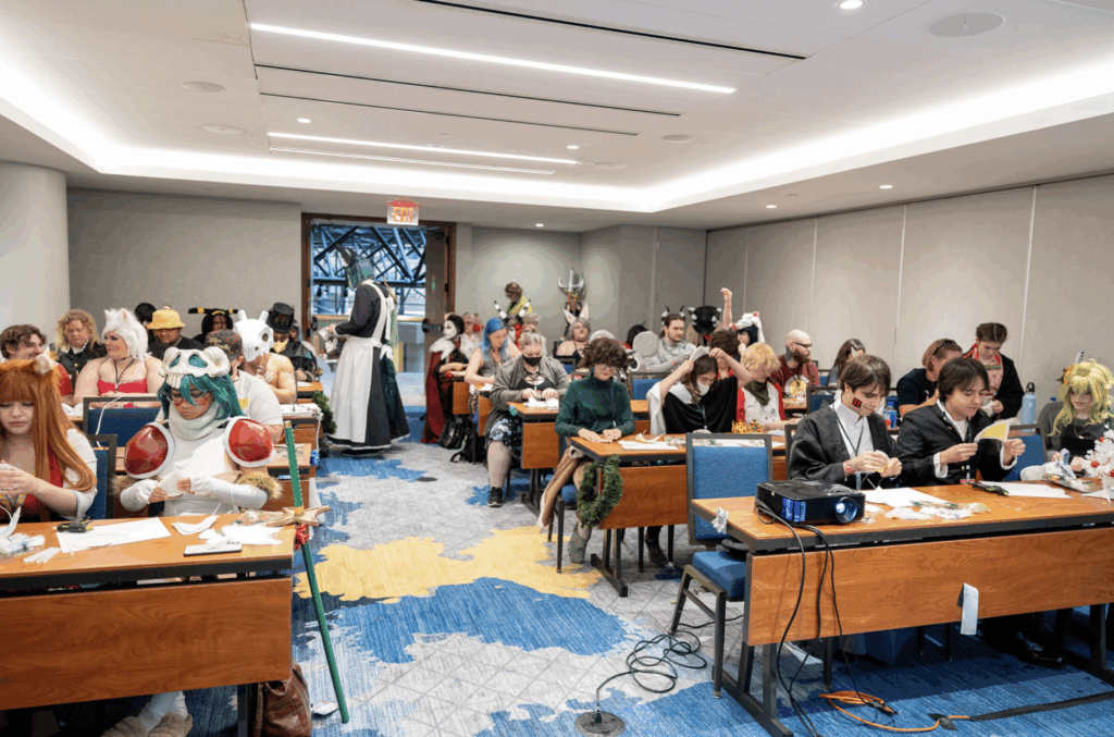 A wide shot of a crowded convention classroom shows numerous cosplayers in colorful and creative costumes seated at desks, working on arts and crafts. Toward the back of the room, Caleb in black-and-white attire with face paint resembling a rabbit stands out, interacting with other attendees. The mood is lively and focused.
