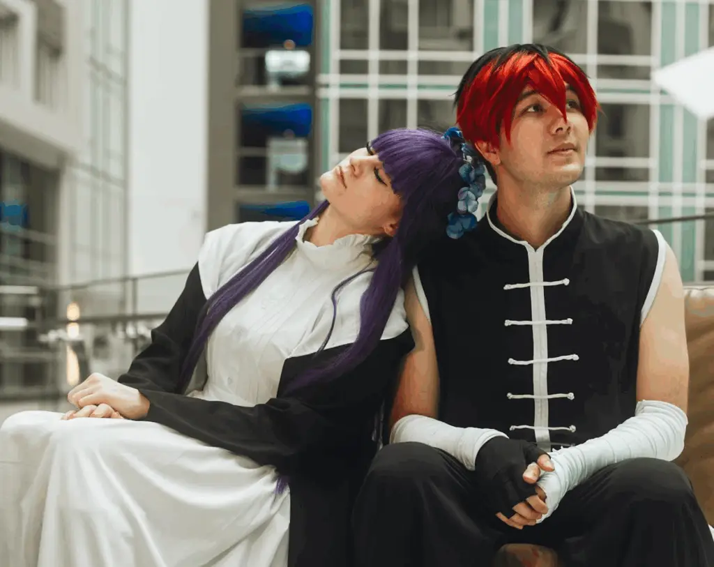Two cosplayers sit closely on a bench in a convention center atrium. The person on the left, with long purple hair and a black-and-white maid-style outfit, rests their head on the shoulder of the person beside them. The person on the right wears a black martial arts-inspired outfit with white trim and red-and-black spiked hair, looking off into the distance. The mood is calm and intimate.