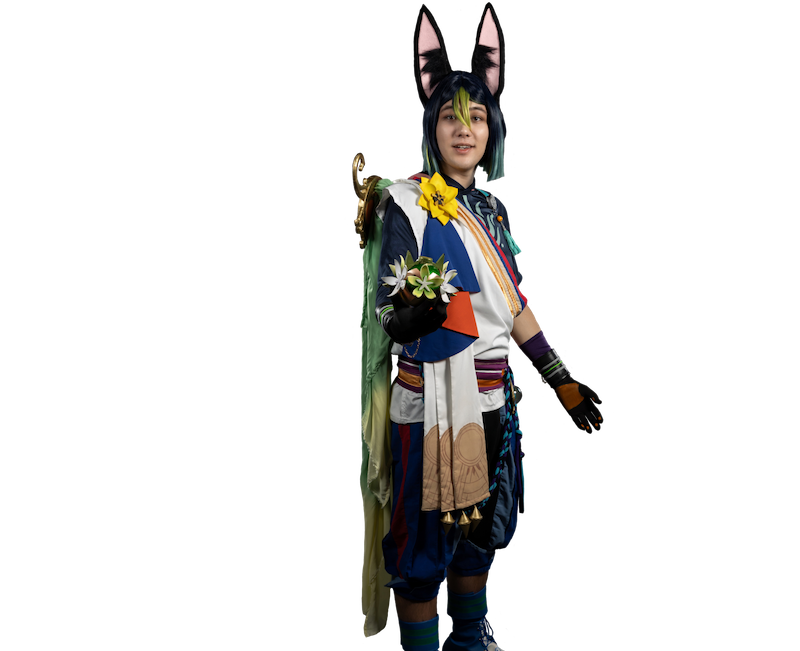 Caleb dressed as a colorful anthropomorphic character stands confidently against a black background. They wear tall black fox ears with pink insides, a blue-and-green wig with lime green bangs, and a richly detailed outfit with vibrant patterns, layered fabrics, and a large yellow flower on their shoulder. The costume includes gloves, sashes, and accessories, with one hand extended holding a small cluster of silver flowers. The expression is welcoming and calm, conveying a sense of nature or magical energy.